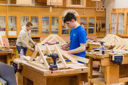 Three individuals working in a woodworking classroom, assembling wooden frames at benches surrounded by tools and materials.