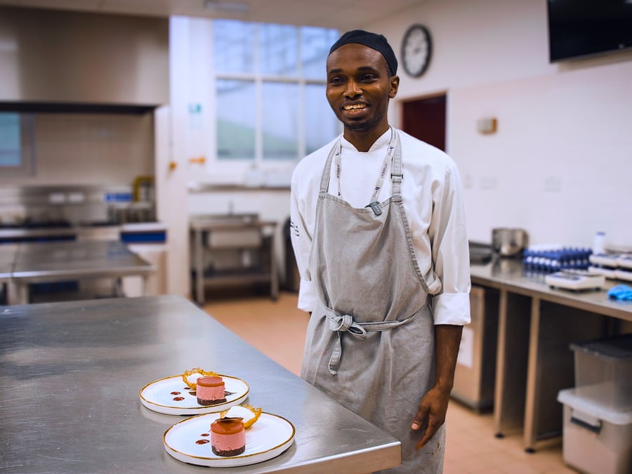 Smiling chef in white coat and apron standing in commercial kitchen with plated desserts