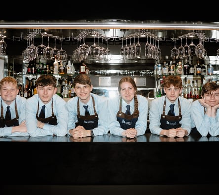 Hospitality students at Highlands College standing behind a bar counter wearing ‘Barista’ aprons, with wine glasses overhead and a stocked beverage shelf in the background.
