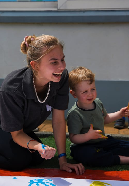 A vertical photo of a student on her knees laughing beside a child who is sat on the floor and looking concerned.