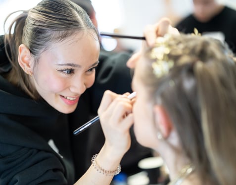 Beauty therapy student at Highlands College applying eye makeup with a brush to a client with styled hair and decorative elements.