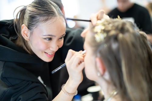 Beauty therapy student at Highlands College applying eye makeup with a brush to a client with styled hair and decorative elements.