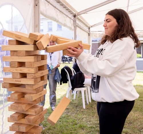 Student playing giant Jenga under a white tent on a grassy area at a Highlands College event, with others watching in the background.
