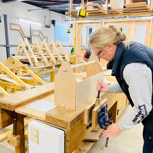 A photo of a woman in a wood workshop using a vice. She has a handmade wooden tool box in front of her on the workbench.