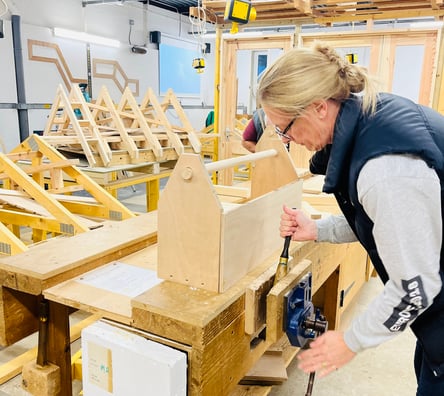 A photo of a woman in a wood workshop using a vice. She has a handmade wooden tool box in front of her on the workbench.