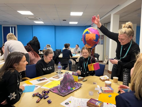 A medium shot of a classroom environment. A group sits at a table covered with chocolates, academic papers, and primary school science projects.