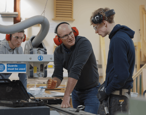 A medium shot of three individuals wearing ear protectors in a workshop. Two individuals are engaged in conversation whilst the third watches from behind industrial machinery.