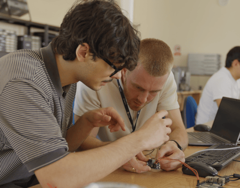 A medium shot of two individuals sat at a desk looking intensely at a small PCB with cables coming out of it.