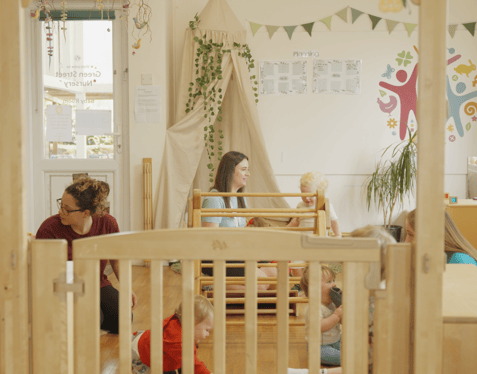 A wide shot of a playroom environment. Six babies play with toys under the supervision of three individuals.